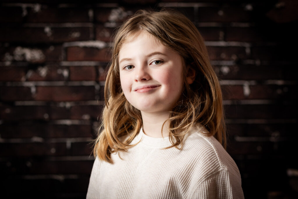 A young boy smirking at the camera in front of a brick wall.