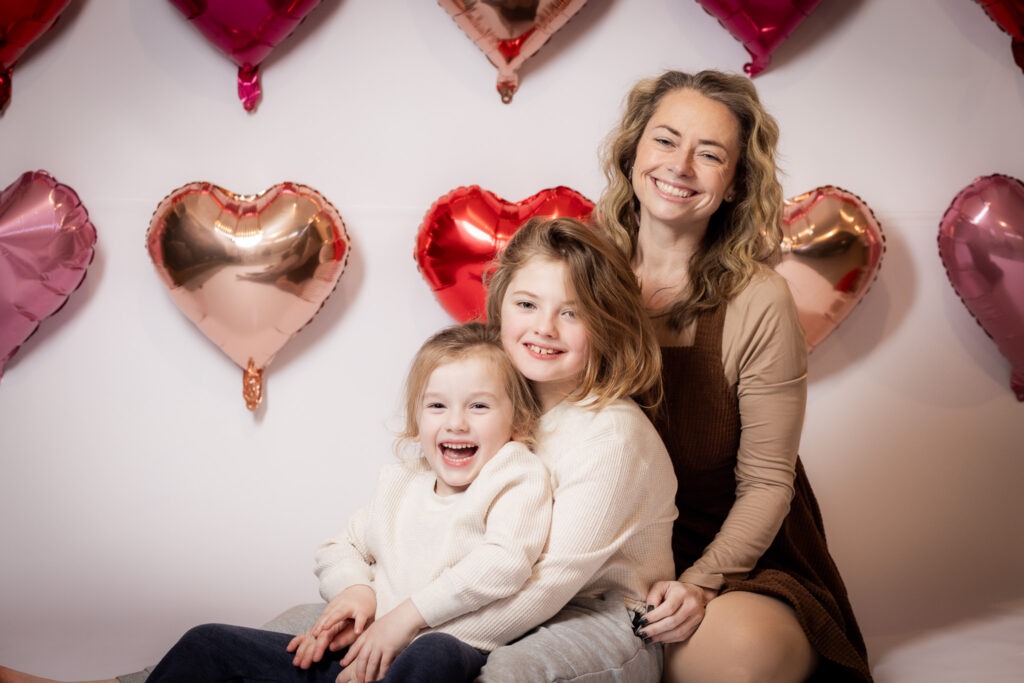 Mom and two young sons smiling in front of a valentines day heart balloon.